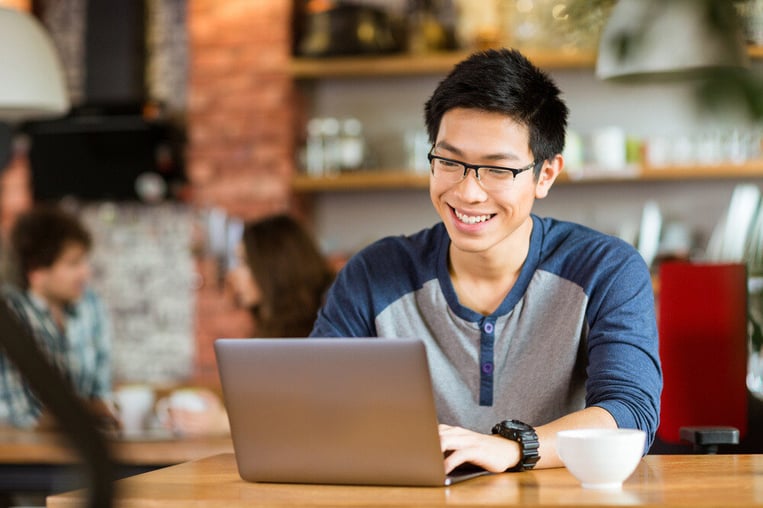 Joven asiático con gafas sonriendo y utilizando un portátil en una cafetería